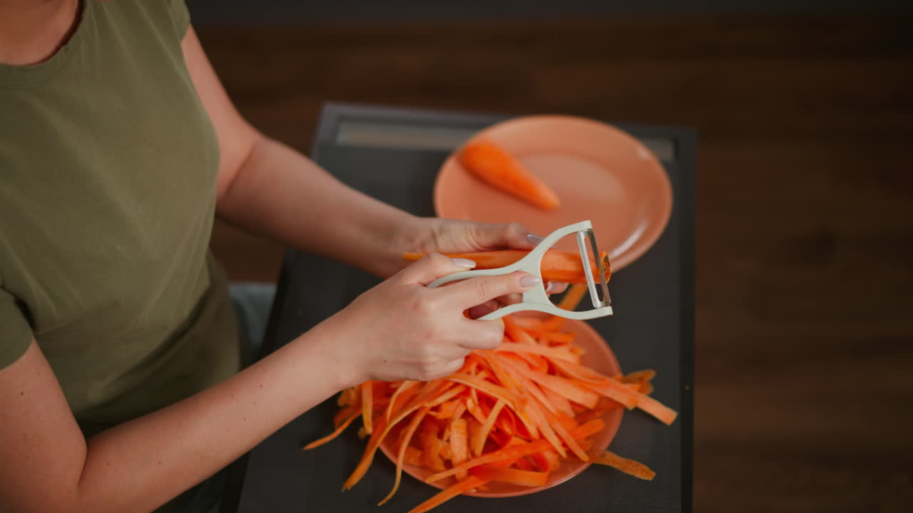 Overhead view of person peeling carrot with white peeler, hand holding orange vegetable above plate filled with long peeled strips, another plate nearby holding single carrot on dark kitchen tabl
