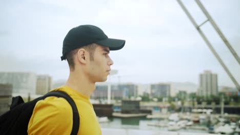 Young-Attractive-Trendy-young-Man-wearing-a-cap-and-a-backpack-walking-on-a-bridge-on-a-sunny-day-with-an-urban-city-background