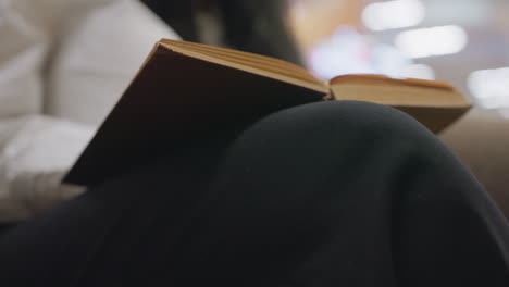 close up showing woman resting open vintage book across crossed leg gently turning pages with manicured fingers while sitting calm lounge chair, blurred warm lights