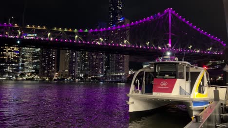 Cityhopper-Atracó-En-La-Terminal-De-Ferry-De-Howard-Smith-Wharves-Esperando-Su-Viaje-Programado-Por-La-Noche-Con-El-Icónico-Puente-De-La-Historia-Iluminado-En-Púrpura,-Reflejado-En-El-Agua-Ondulada-Del-Río-Y-El-Paisaje-Urbano-Del-Centro-Urbano