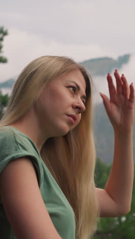 attractive young woman brushes long hair standing against old mountains covered with descending white misty clouds at resort slow motion