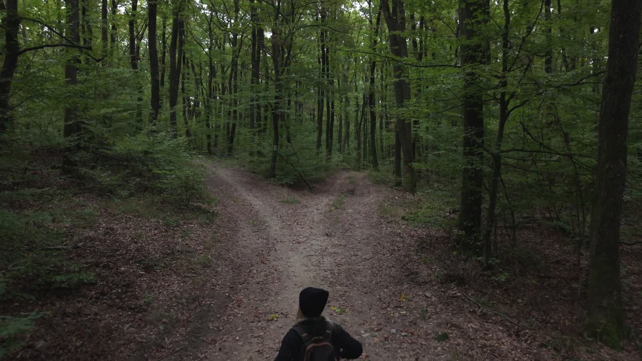 A woman with a backpack walks along a dirt path deep in a dense green forest, approaching a fork in the trail. The peaceful atmosphere and natural surroundings evoke solitude and exploration