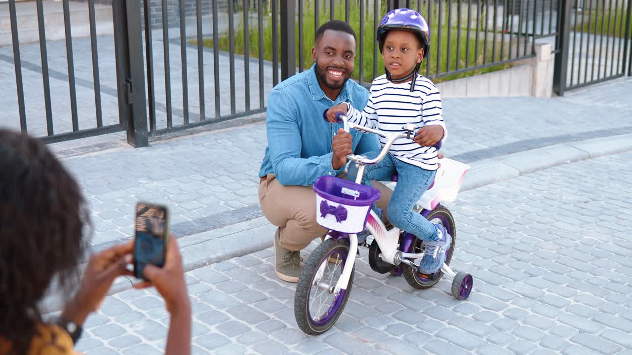 Back view on African American mother taking photo with smartphone camera of husband and daughter on bike