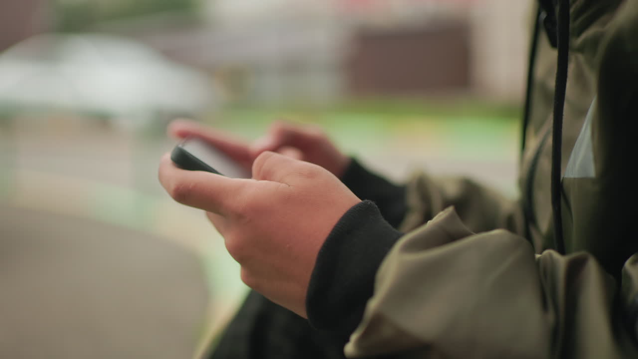 Close up of hands holding handheld device as boy in jacket plays game outdoors, blurred greenery and cars in background highlighting focus on digital interaction and modern youth lifestyle