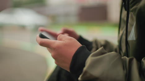 close up of hands holding handheld device as boy in jacket plays game outdoors, blurred greenery and cars in background highlighting focus on digital interaction and modern youth lifestyle