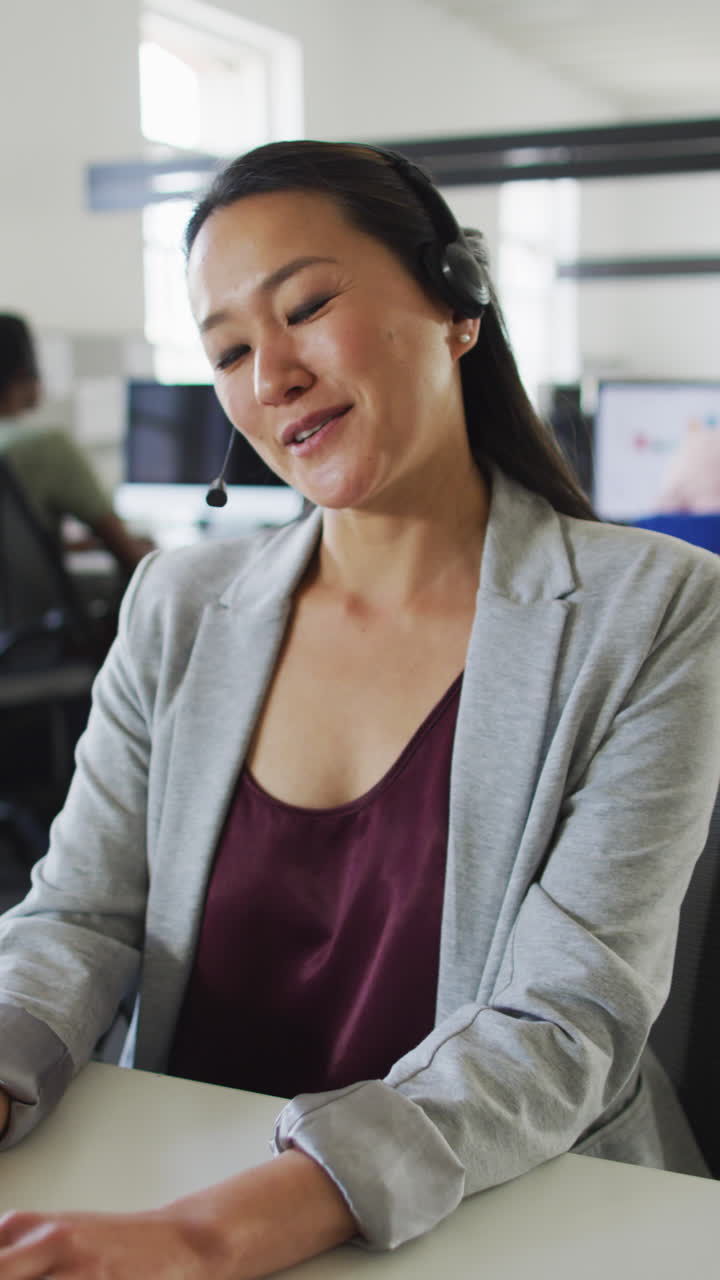 video vertical de una mujer asiática sonriente sentada en un escritorio hablando por auriculares en la oficina