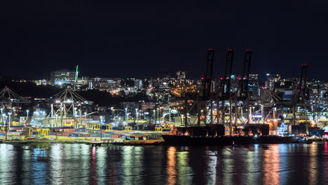 Auckland,-Puerto-De-Nueva-Zelanda,-Lapso-De-Tiempo-Nocturno-Con-Luces-Reflejándose-En-El-Agua-Mientras-Los-Transportistas-Apilan-Contenedores-De-Envío