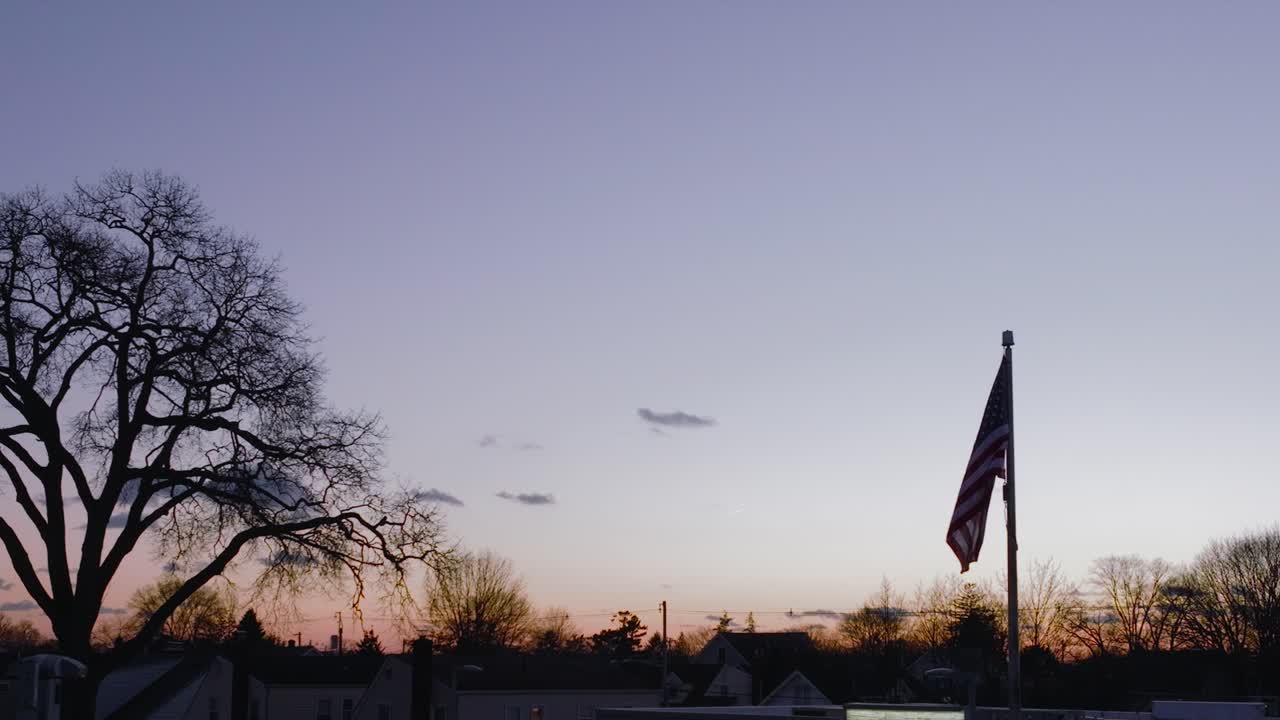 Sebuah bidikan udara, pada ketinggian rendah, bendera Amerika berkibar tertiup angin saat matahari terbenam yang indah, dalam gerakan lambat dengan siluet pepohonan di latar belakang.