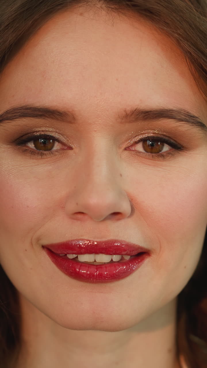 Happy woman with bright red lip gloss looks in camera smiling. Portrait of young model with professional makeup on blurred background closeup