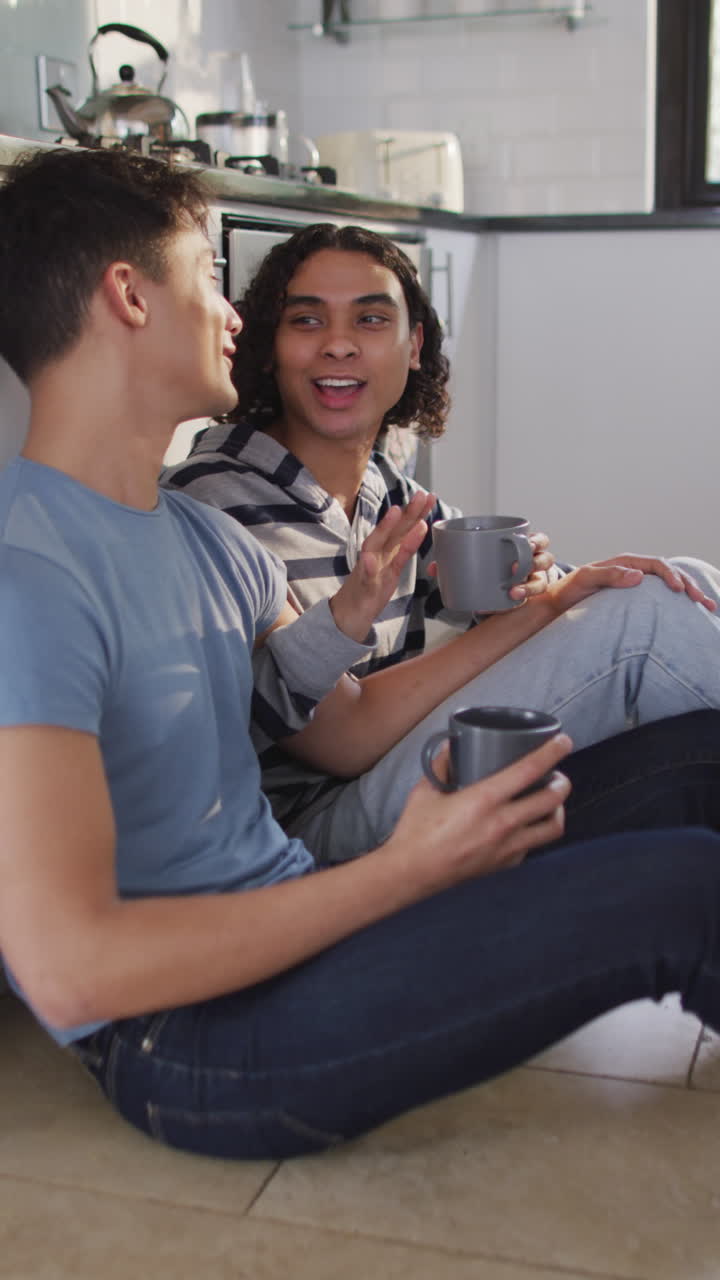 video vertical de una pareja feliz y diversa sentada en el suelo y bebiendo café en la cocina