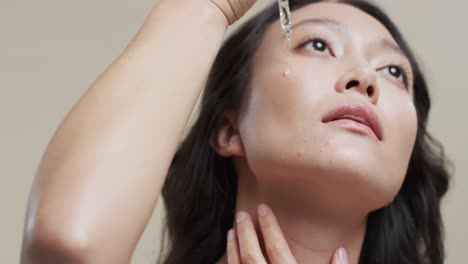 어두운 머리카락을 가진 아시아 여성이 베이지색 배경에 얼굴에 화장품을 적용하고, 느린 움직임
