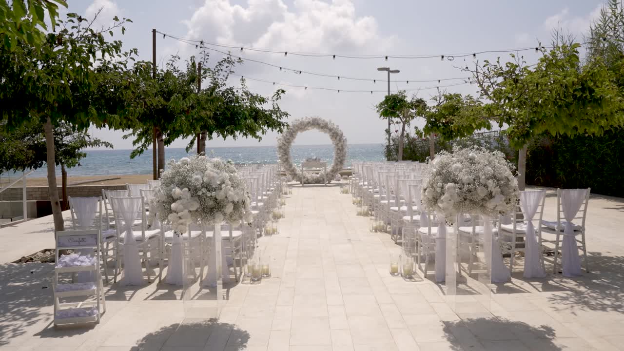 This serene beachside location is arranged for a wedding ceremony. White chairs, floral decorations, and an arch create a romantic atmosphere on a sunny day