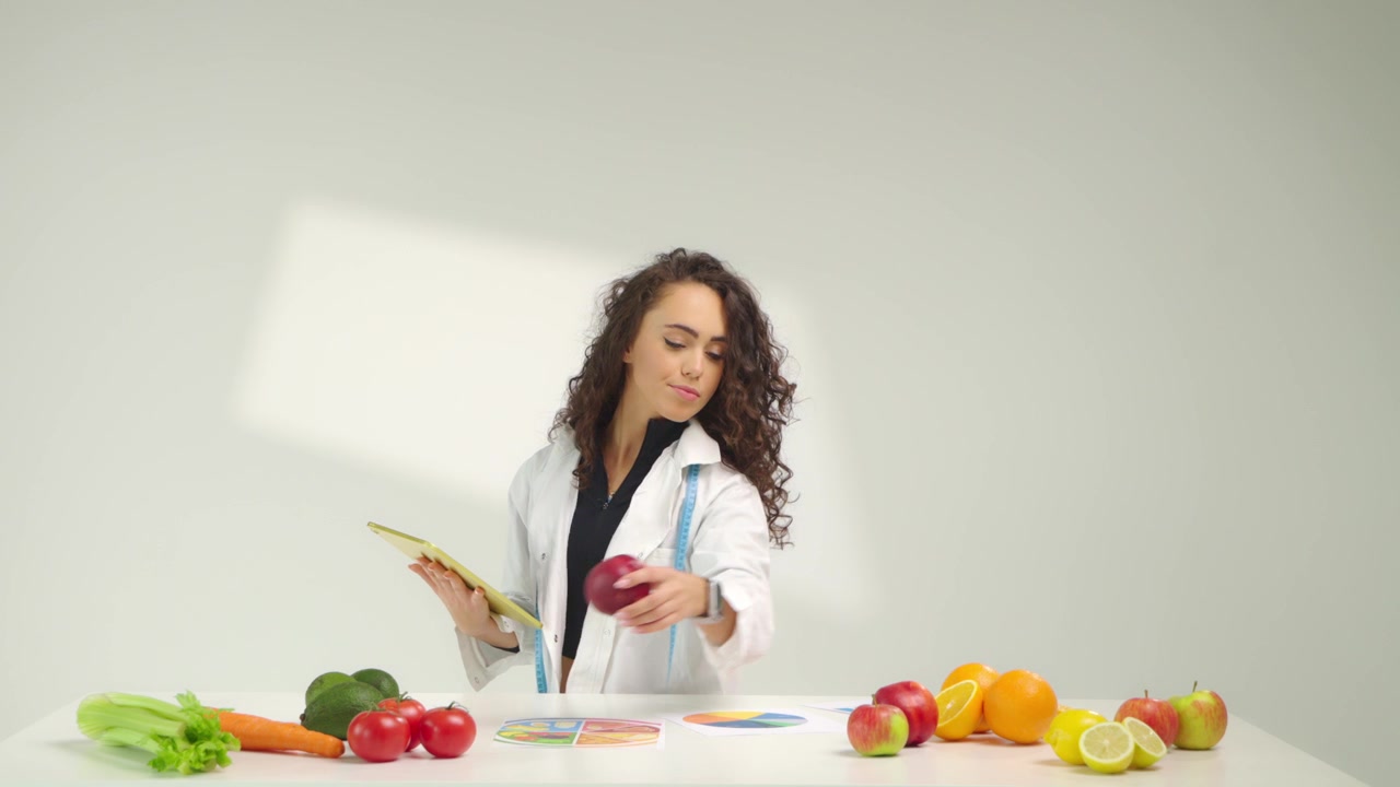 Specialista nutrizionista donna concentrata che lavora con un tablet mentre sistema verdura e frutta su un tavolo in uno studio bianco. Concetto di pianificazione della dieta, stile di vita sano e consulenza sul benessere.