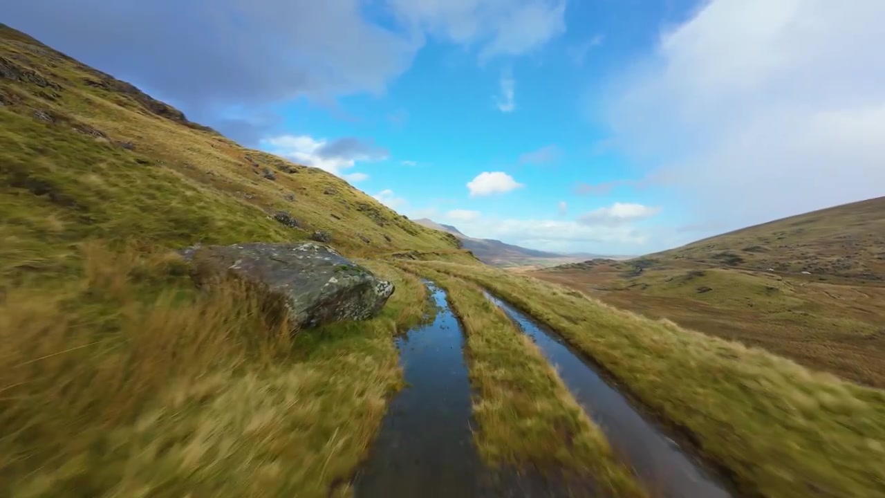 First person perspective journeying along a waterlogged, rocky footpath through a vast, grassy mountain valley under a bright blue sky with scattered clouds, simulating a dynamic outdoor adventure