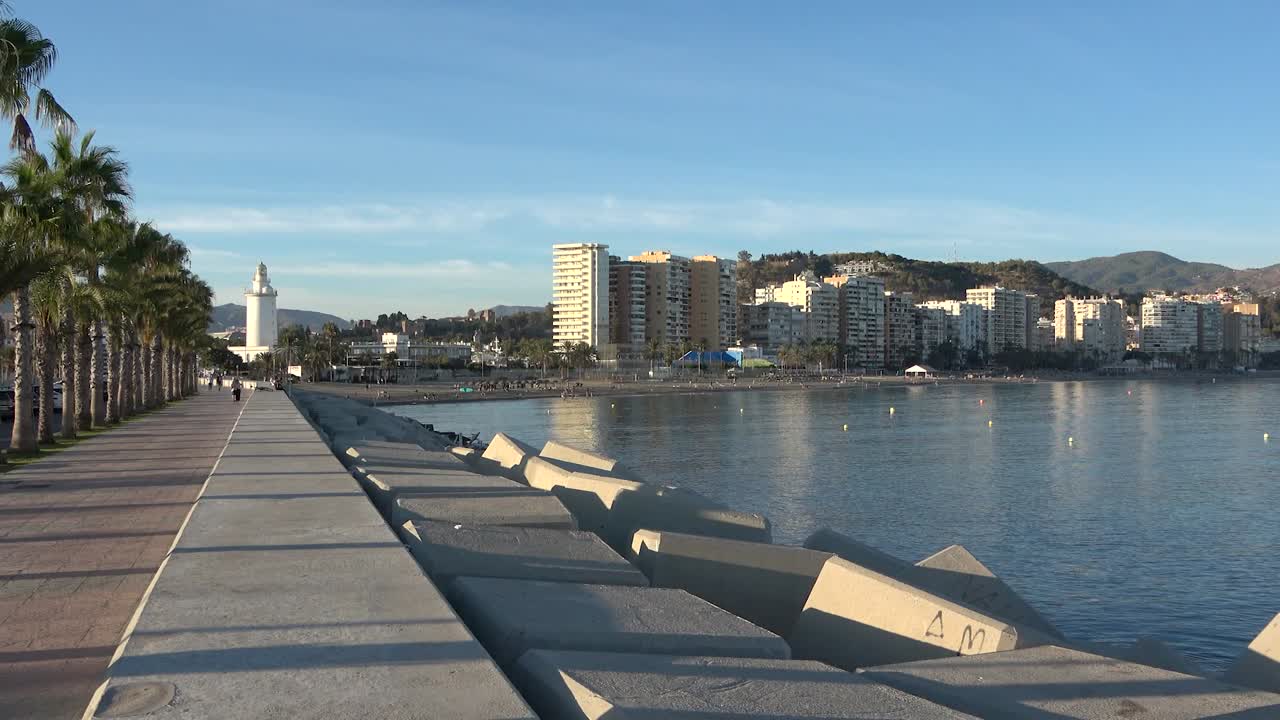 Frangiflutti in cemento lungo la passeggiata con mare calmo, faro in lontananza, edifici costieri e montagne sullo sfondo che creano un'atmosfera rilassante da costa mediterranea durante il giorno
