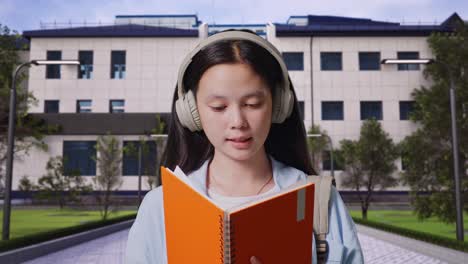 close up of asian teen girl student with a backpack holding and reading a book while standing in front of a school building