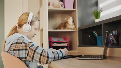 a girl in headphones draws on a graphics tablet in her room. in the background, her cat is sitting on a shelf.