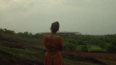 A-woman-stands-in-a-field-gazing-at-a-distant-building-under-construction-on-a-cloudy-day