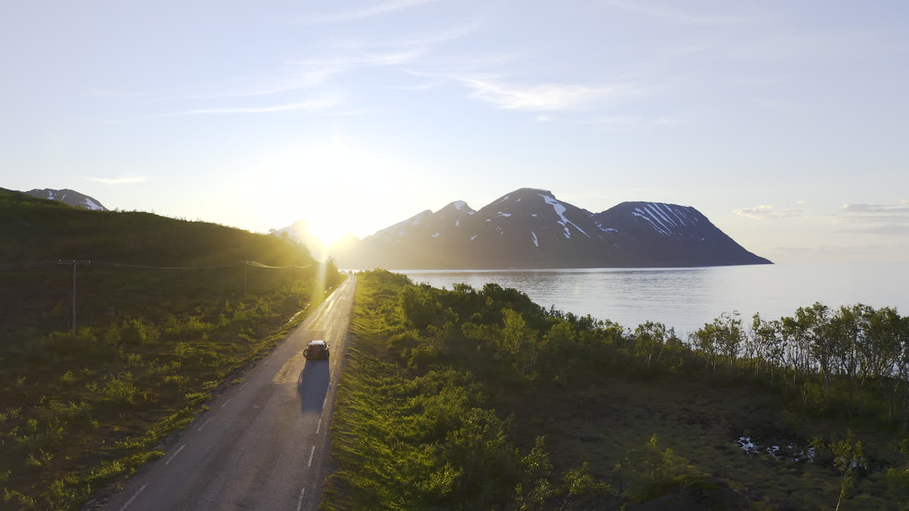 coche conduciendo una pequeña carretera hacia la puesta de sol junto a un océano y un cielo azul con un paisaje montañoso en el fondo