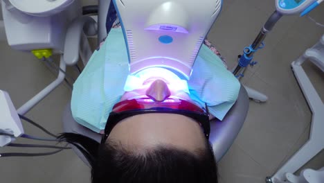 young woman with an expander in mouth and red protective glasses getting uv whitening at the dentist's office by an ultra violet machine.