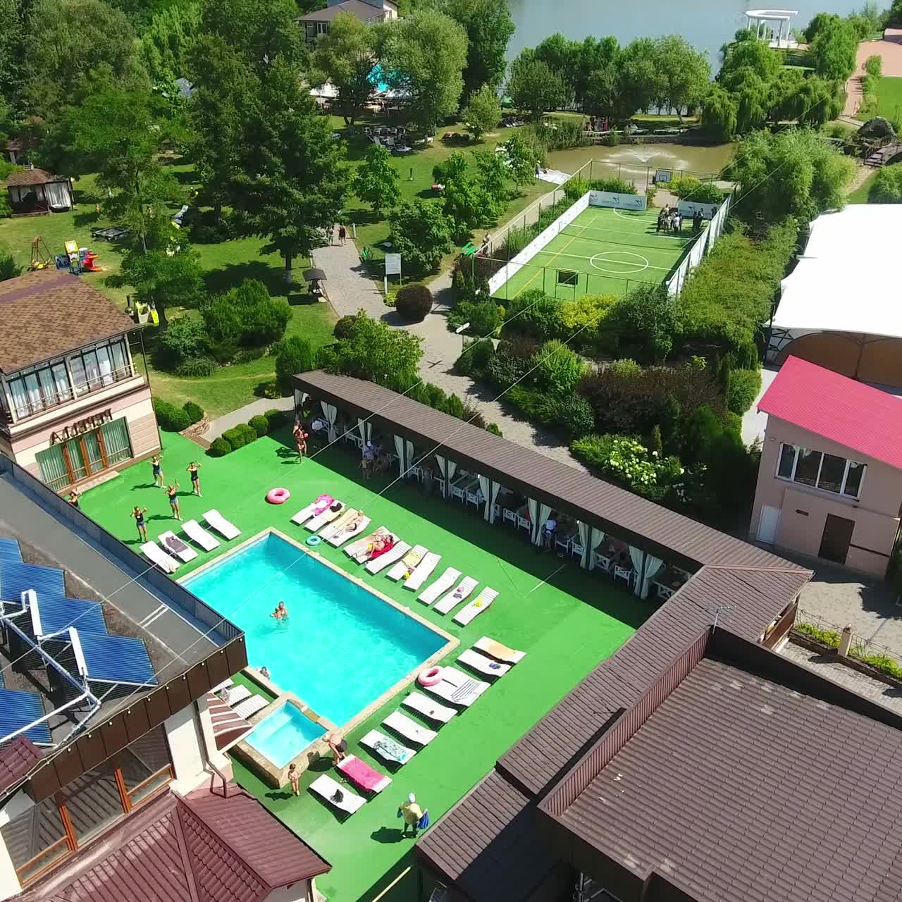 People resting at spa recreational complex in rural area. Green beautiful territory with swimming-pool, tennis court and pond with fountain. Drone footage