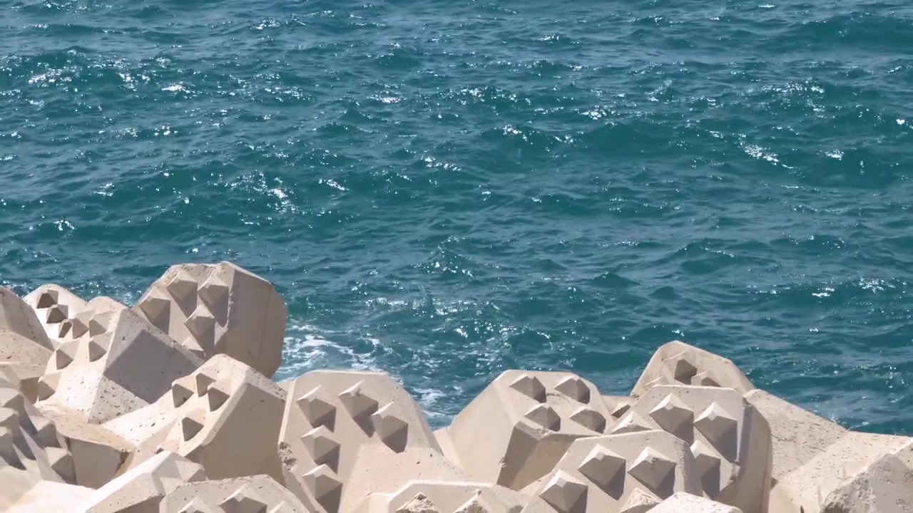 Statisk vidvinkelbillede af blå havbølger, der slår mod kystbeskyttelsesstrukturer af beton-tetrapoder.