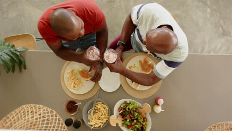 eating burgers and fries, african american father and adult son enjoying lunch together at home