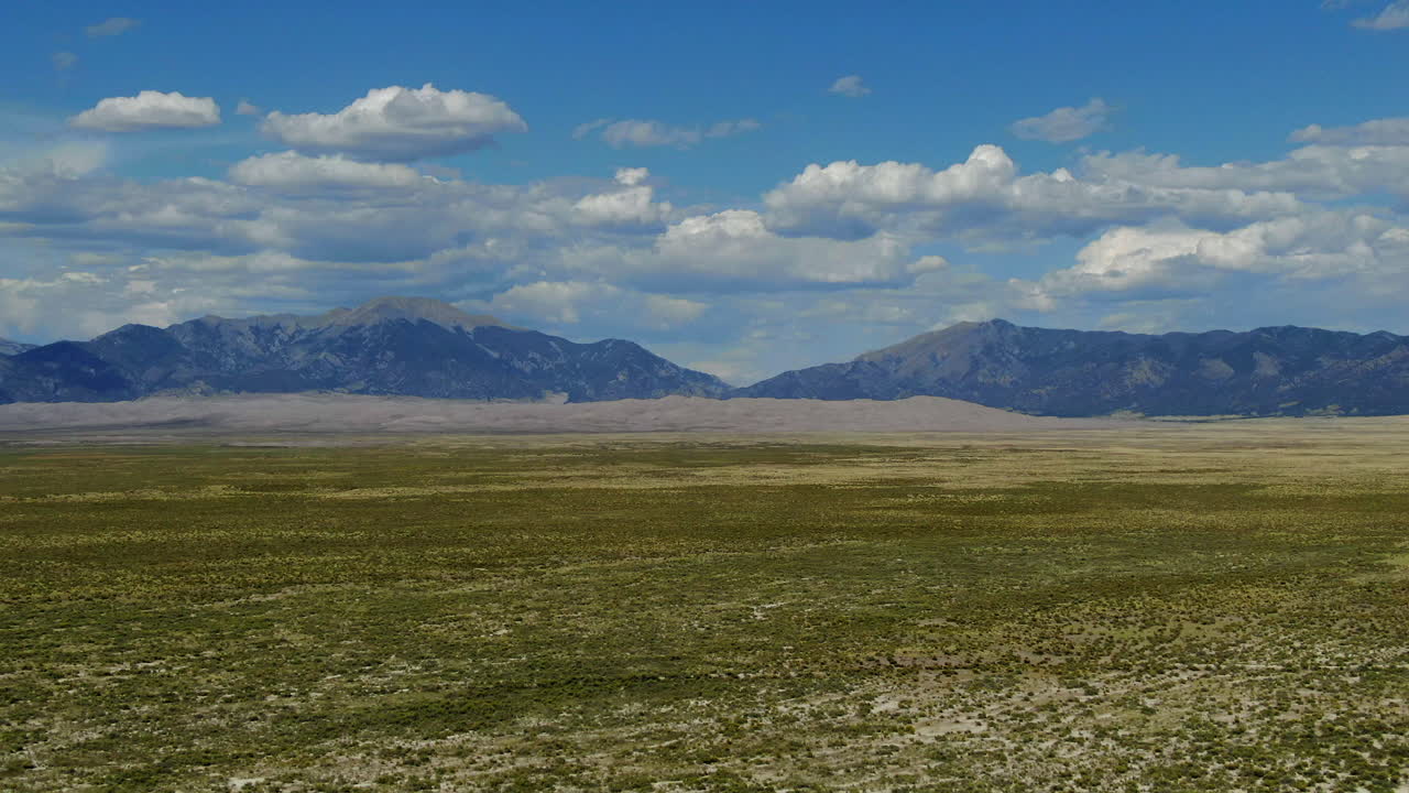 Filmatisk droneoptagelse fra luften i sensommeren af indgangspartiet til Great Sand Dunes National Park, Colorados Rocky Mountains' 14er-toppe, med sprødt, gyldentgult højt græs, blå himmel, skyer og fremadgående bevægelse