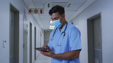 Mixed-race-male-doctor-wearing-face-mask-standing-in-hospital-corridor-using-tablet