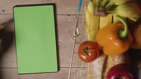 overhead studio shot of person using green screen digital tablet next to basic food items in supermarket wire shopping basket 1