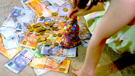 la niña toma monedas en la parte superior de los billetes sudafricanos y los depósitos en la caja de dinero