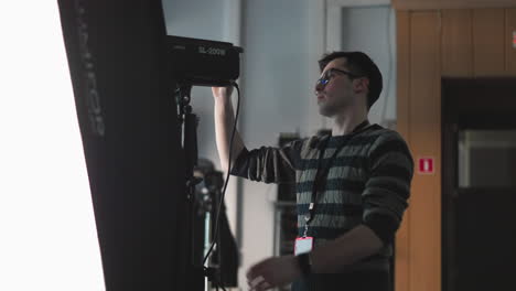 camera technician adjusting power switch and brightness knob on sl200w led studio light mounted on tripod bracket with cables visible against diffuser and control rack in broadcast production set