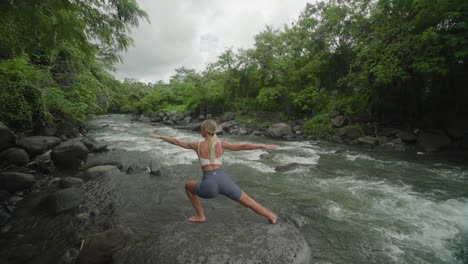 woman practicing warrior pose on rock along calming flow of water in jungle