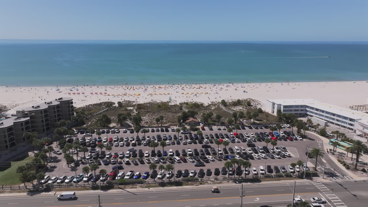Aerial view of Saint Pete Beach public parking lot packed with cars, beach access boardwalk, beachgoers with umbrellas, and stunning turquoise Gulf waters in Florida's popular beach destination