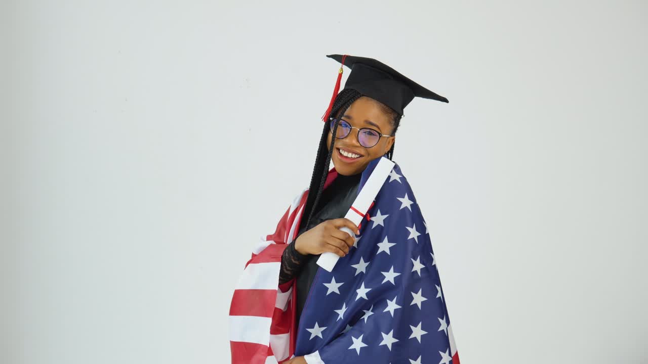 feliz elegante mujer afroamericana estudiante en uniforme de posgrado muestra diploma sosteniendo bandera de ee.uu. en los hombros. educación de ee.