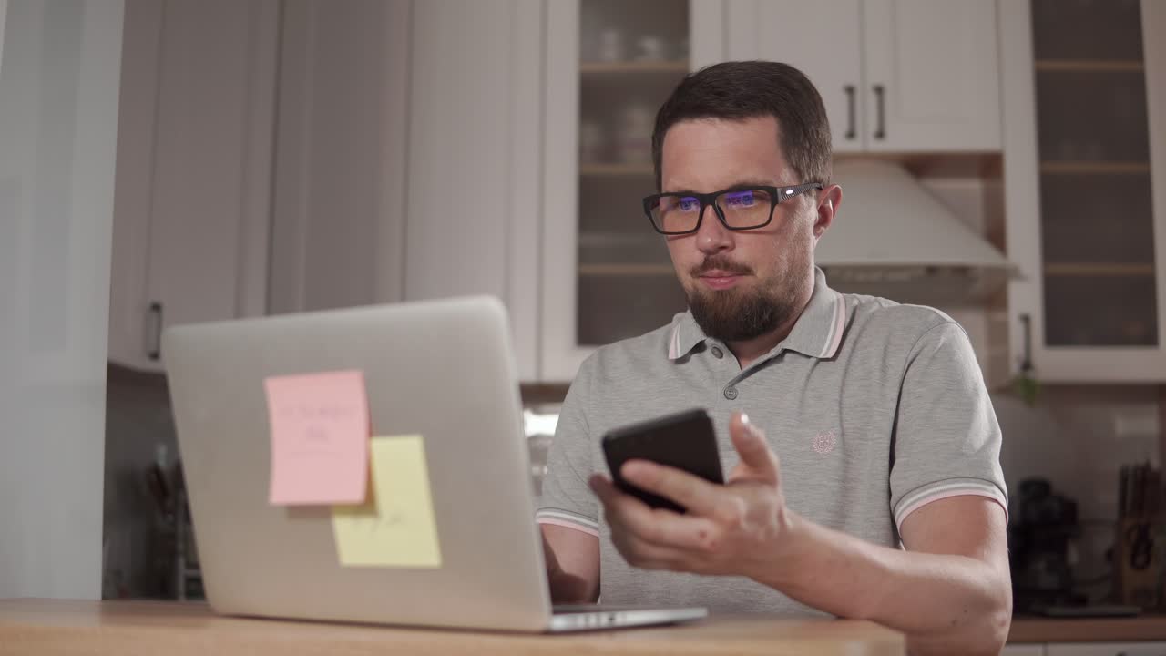 hombre trabajando desde casa, cansado mirando el teléfono y la computadora portátil