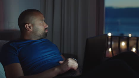 young man using laptop lying in bed browsing online finished working late at night looking at watch