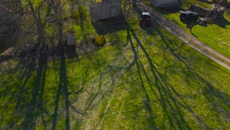 aerial drone footage following a small retro and vintage tractor working in a sunny green grassy field during summer time at a countryside garden or a yard that has large trees that cast shadows on it