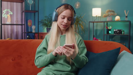 Happy-caucasian-woman-in-wireless-headphones-relaxing-sits-on-sofa-at-home-listening-favorite-music