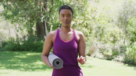 Portrait-of-biracial-woman-holding-yoga-mat-laughing-outside-in-garden-on-a-sunny-day