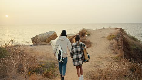 A-happy-brunette-guy-in-a-black-hat-in-a-white-sweatshirt-and-his-blonde-girlfriend-in-a-checkered-shirt-walk-down-the-slope-to-the-sea-and-hold-surfboards-in-their-hands-near-the-sea-in-sunny-weather-in-the-morning-in-autumn