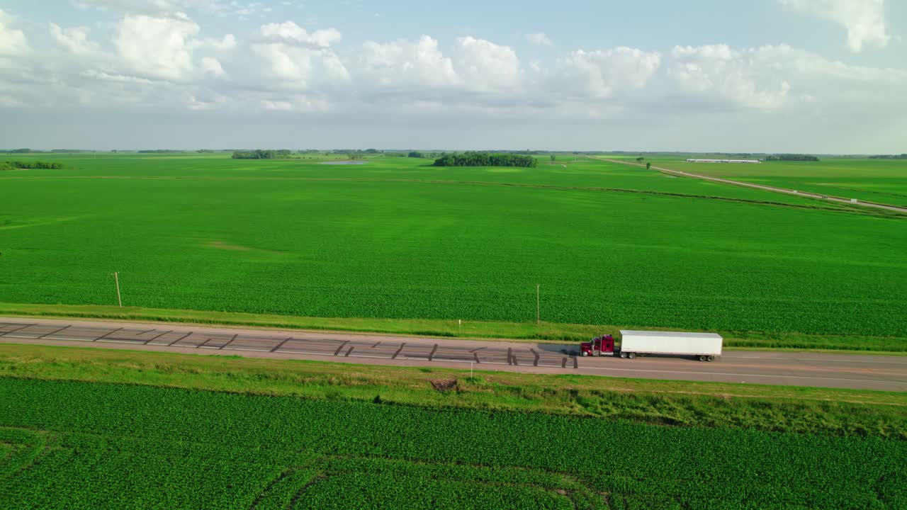 Aerial view of a red semi-truck with a reefer trailer driving along a rural highway through expansive green farmland in Minnesota.