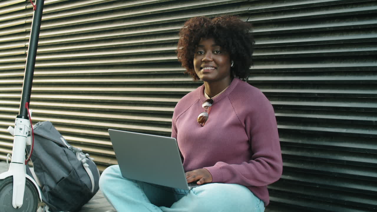 Portrait of Cheerful African American Woman Sitting on Street and Using Laptop