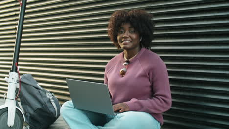 Portrait-of-Cheerful-African-American-Woman-Sitting-on-Street-and-Using-Laptop