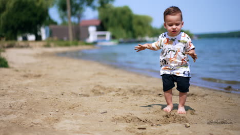 one year old toddler boy plays with sand on the beach. child stands up and looks around holding dirty hands up.