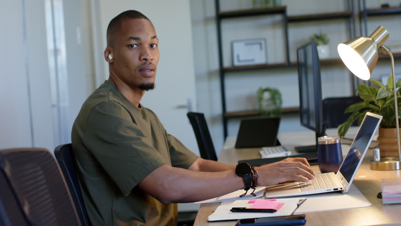 Man typing on laptop at office desk with earbuds and coffee mug pausing while acknowledging camera