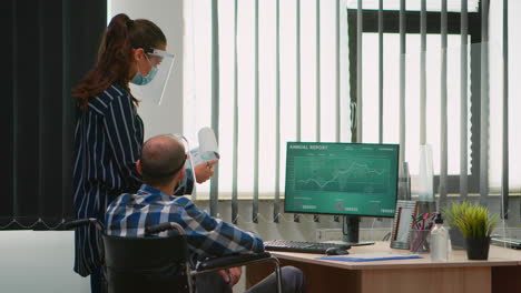 Frau-Mit-Maske-Zeigt-Auf-Den-Desktop-Und-Spricht-Mit-Einem-Behinderten-Kollegen