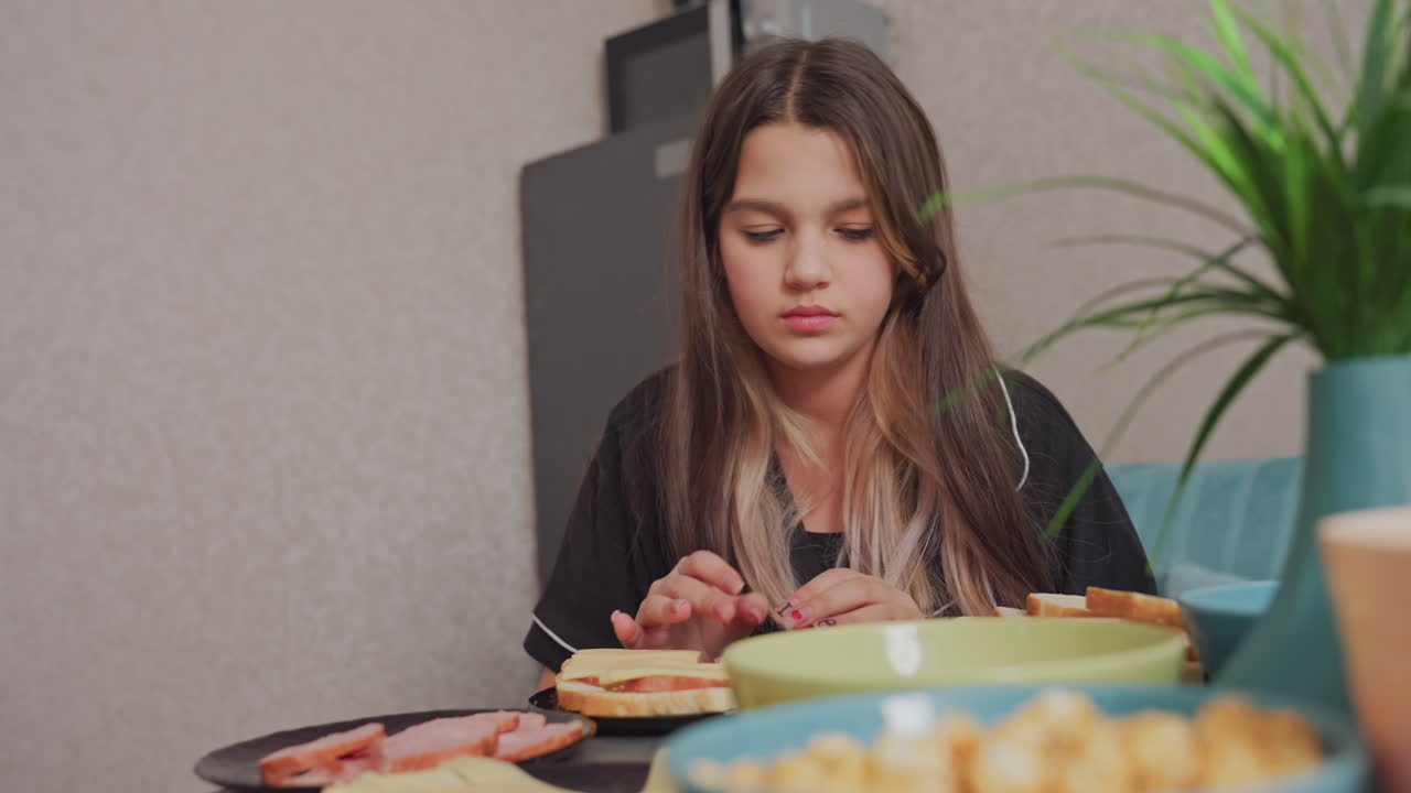 young woman in dark pajamas sits at table preparing breakfast using sliced cheese and sausage, surrounded by bowls and bread in cozy kitchen setting, focused on arranging ingredients with care