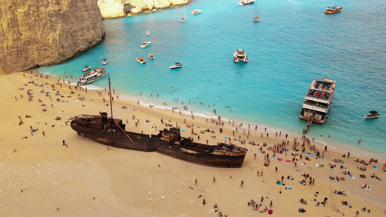 Aerial drone view of the Navagio beach on the Ionian Sea coast of Zakynthos, Greece. Moored boats and resting people, rusty ship, blue water. Slow motion