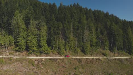 red jeep suv off road vehicle driving on dirt logging road kicking up dust wide aerial angle reversing pan right revealing scenery dense green forest snow covered mountain peaks in wilderness